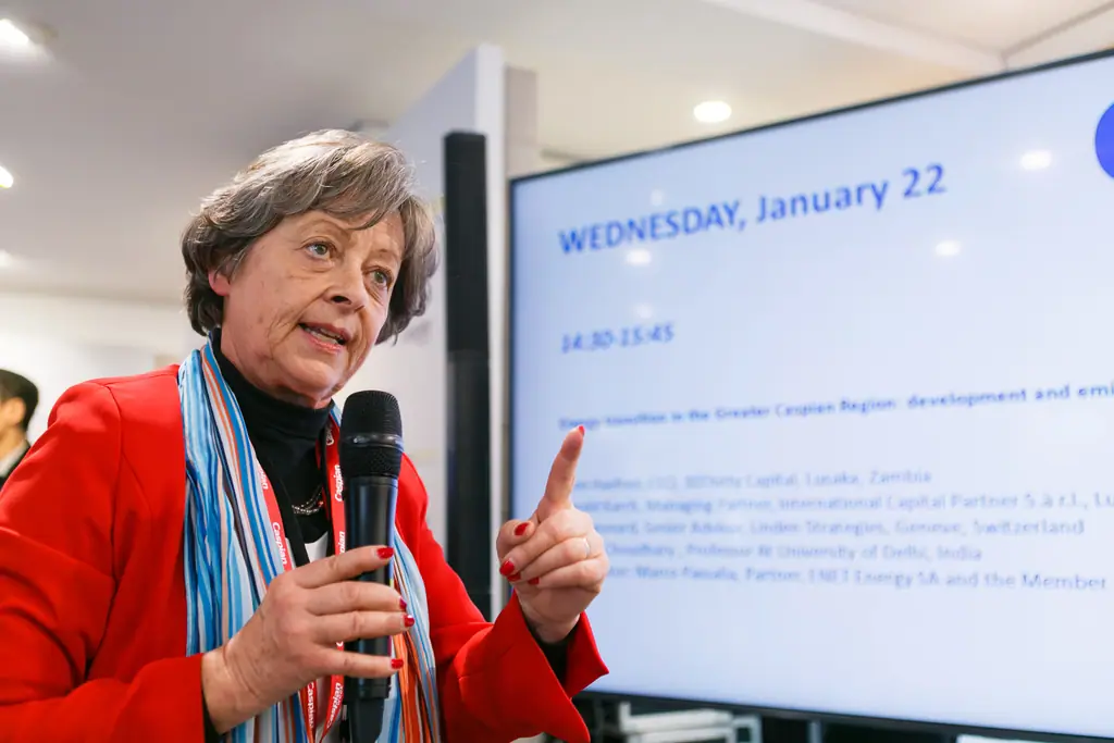 A speaker in a red jacket makes a point, microphone in one hand, the session schedule on a screen behind her